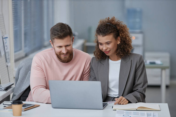 Dos jóvenes economistas en ropa de casualwear inteligente mirando a la pantalla del ordenador portátil mientras están sentados junto al escritorio en la reunión de trabajo y discutiendo datos en línea - Foto, imagen