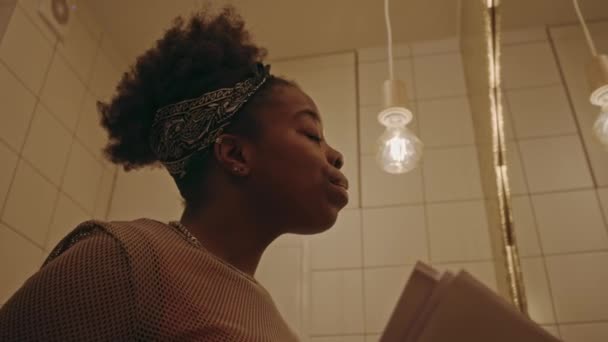 Low angle shot of young African American woman revising study materials on paper sheer while using lash curler, getting ready for exam at university in front of mirror in dimly lit bathroom - Footage, Video