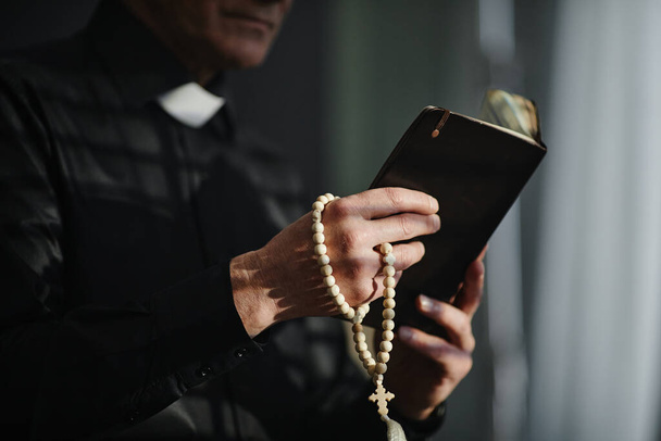 Πλευρική άποψη closeup αγνώριστη ιερέας κρατώντας Αγία Γραφή και κομπολόι, ενώ προσεύχεται στο δραματικό φως, αντίγραφο χώρο - Φωτογραφία, εικόνα