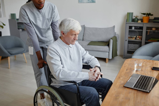 Retrato de vista lateral de hombre mayor de pelo blanco en silla de ruedas en casa con la asistencia de la enfermera - Foto, Imagen