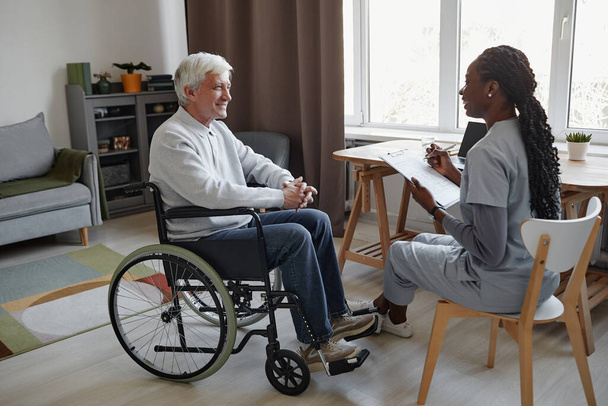 Pełna długość boku widok portret siwowłosy senior z niepełnosprawnością rozmawia z uśmiechniętą pielęgniarką w domu spokojnej starości, przestrzeń kopiowania - Zdjęcie, obraz