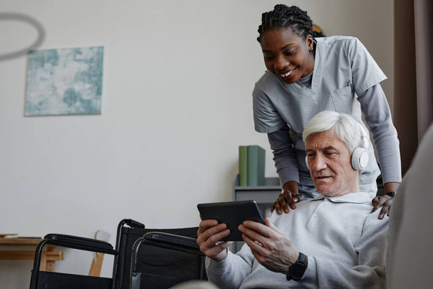 Retrato de hombre mayor de cabello blanco con discapacidad relajándose en casa y usando tableta con enfermera asistida, espacio para copiar - Foto, Imagen