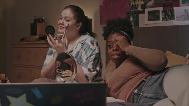 Dos jóvenes bastante multiétnicas compañeras de piso chateando mientras hacen maquillaje, sentadas en la cama en un acogedor dormitorio y preparándose para salir por la noche - Imágenes, Vídeo
