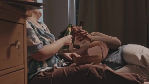 Две многоэтнические веселые девушки лежат на кровати с бутылками сидра и сплетничают во время ночевки - Кадры, видео