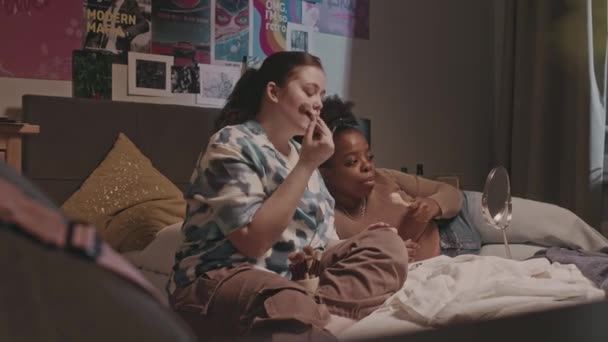 Dos jóvenes diversas novias charlando y haciendo maquillaje en la cama en el acogedor dormitorio durante la pijamada - Metraje, vídeo
