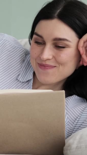 Hymyilevä valkoihoinen nainen lepää sohvalla ja lukee kirjaa kotona. Rentouttava ja viettää viikonlopun vapaa-aikaa suosikki harrastus. Pystysuora video. - Materiaali, video