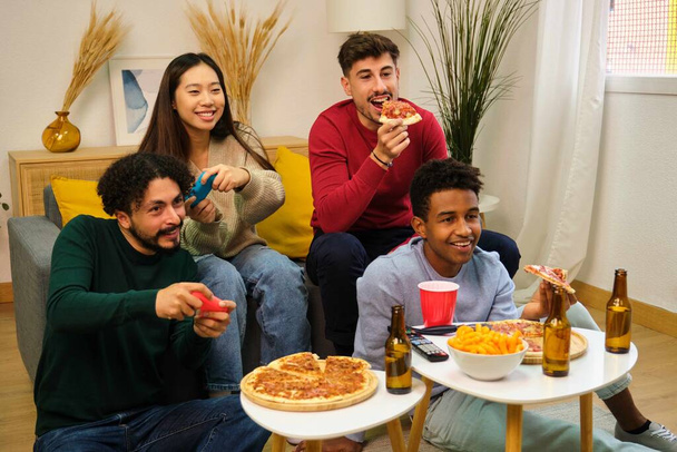 Группа многорасовых друзей, которые едят пиццу и играют в видеоигры в общей квартире. - Фото, изображение