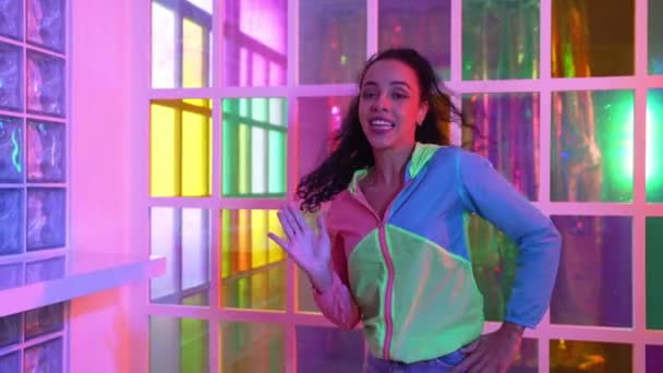 Happy hispanic girl in lively mood moving along music in neon light while wearing colorful cloth at night club. Street dancer making energetic footstep or movement while looking at camera. Regalement. - Footage, Video