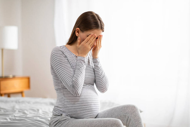 Haavoittuva eurooppalainen raskaana oleva nainen sängyllä peittää kasvonsa, koskettava kuvaus kodin lohdutuksesta raskauden aikana emotionaalisia hetkiä. - Valokuva, kuva