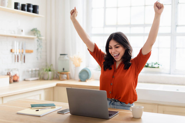 Молодая женщина за кухонным столом выражает волнение с поднятыми руками и открытым ноутбуком, работая в Интернете - Фото, изображение