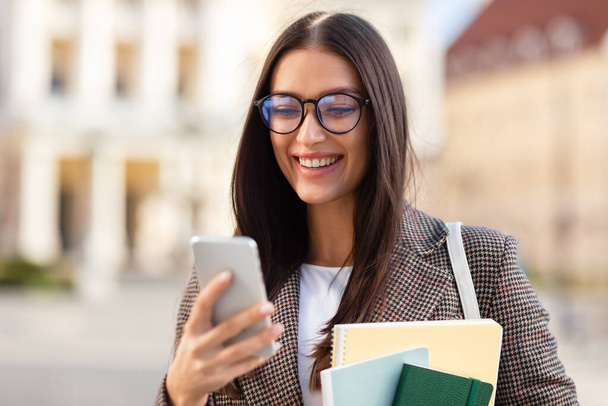 Nahaufnahme einer jungen Frau in stylischer Kleidung, die mit einem Lächeln auf ihrem Handy surft und sich auf Konnektivität und Technologie konzentriert - Foto, Bild
