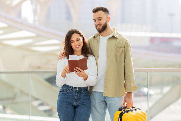 Un jeune couple souriant debout dans un aéroport, tenant un passeport et une valise jaune, heureux époux millénaires prêts pour les voyages de vacances, vérifier les documents - Photo, image