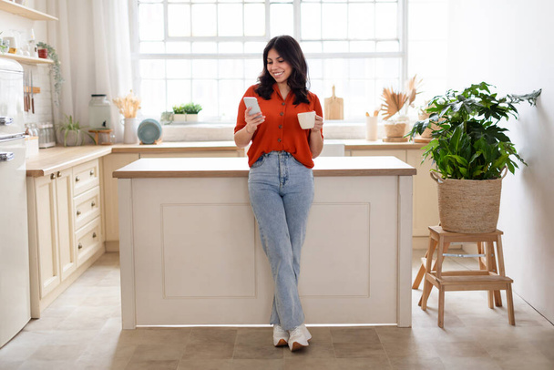 Entspannte junge Frau steht in einer gut beleuchteten Küche mit Kaffeebecher und Smartphone in der Hand und stöbert morgens - Foto, Bild