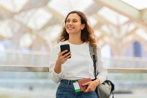 Χαρούμενη νεαρή γυναίκα που κατέχει διαβατήριο και smartphone, ενώ περπατάτε στο αεροδρόμιο, ευτυχισμένη χιλιετή γυναίκα που ταξιδεύει μόνη της, ενθουσιασμένος για το επερχόμενο ταξίδι, αντίγραφο χώρο - Φωτογραφία, εικόνα