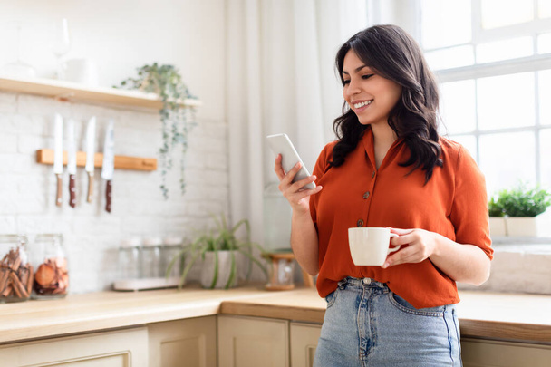 Χαμογελώντας νεαρή γυναίκα απολαμβάνοντας τον καφέ της, ενώ χρησιμοποιεί το smartphone της σε ένα σπιτικό περιβάλλον κουζίνας, αντιγραφή χώρου - Φωτογραφία, εικόνα