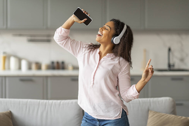 Eine temperamentvolle Afroamerikanerin tanzt frei in ihrer heimischen Küche und hält ihr Smartphone als Mikrofon - Foto, Bild