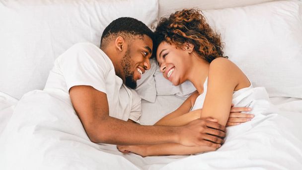 Bom dia, luz do sol Visão superior sobre jovem casal negro amoroso deitado na cama e olhando um para o outro com sorriso, panorama
 - Foto, Imagem