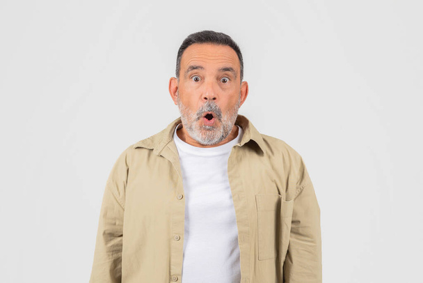 Un hombre de mediana edad con barba gris muestra una expresión sorprendida, con los ojos abiertos y la boca abierta sobre un fondo gris - Foto, imagen