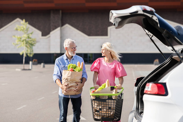 Egy idős, nyugdíjas pár megpakolja az autóját egy bevásárlókirándulásról származó élelmiszerekkel, bemutatva a független, aktív életmódjukat. - Fotó, kép