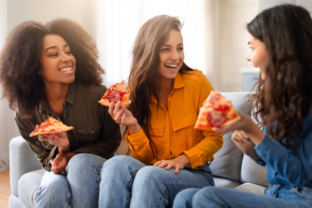 Drie jonge multiraciale vriendinnen lachen en delen pizza in een gezellige huiselijke setting, die warmte en gezelligheid uitstralen - Foto, afbeelding