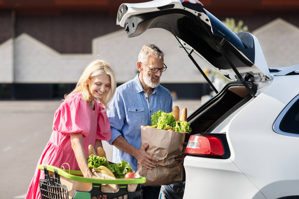 Esta imagen captura a una pareja de ancianos casados mientras cargan comestibles en su auto, ilustrando un recado rutinario pero alegre - Foto, Imagen