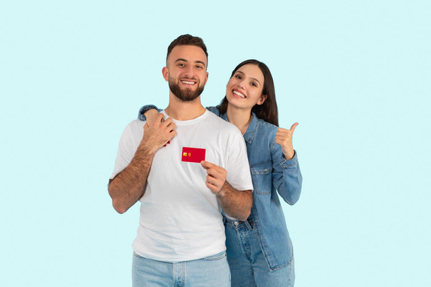 Een vrolijk jong stel poseert met een creditcard, de vrouw geeft een duim omhoog, geïsoleerd op een blauwe achtergrond - Foto, afbeelding