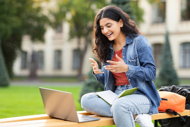 Freudestrahlend lacht eine Inderin, während sie auf einer Campus-Bank auf ihren Laptop blickt und das pädagogische Engagement und den lebhaften Geist eines östlichen Zoomers in einem akademischen Umfeld hervorhebt. - Foto, Bild