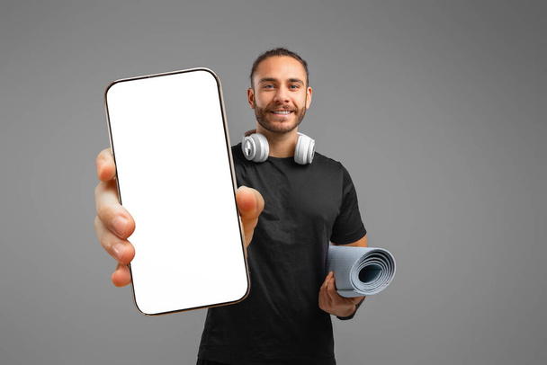 Hombre que presenta una pantalla de teléfono inteligente en blanco mientras sostiene una estera de yoga, lo que indica un anuncio o una promoción de la aplicación - Foto, imagen