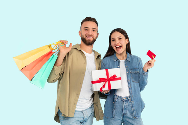 Renkli alışveriş çantaları taşıyan neşeli genç bir çift ve açık mavi arka planda bir kredi kartı sunan bir hediye kutusu. - Fotoğraf, Görsel