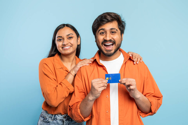 クレジットカードを持っているオレンジ色のシャツの陽気なインディアンカップル, 青のショッピングや支払いを示唆 - 写真・画像
