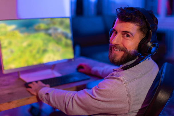 Sonriente joven barbudo jugador hombre admira vívidos gráficos del juego en su monitor de ordenador en una habitación iluminada por neón - Foto, imagen