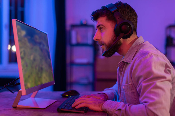 Em uma sala iluminada por luz de néon suave, um jogador cara joga um jogo de tiro em primeira pessoa em sua configuração moderna do PC - Foto, Imagem