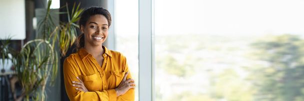 Πορτρέτο της αυτοπεποίθηση αφροαμερικανός επιχειρηματίας στέκεται με διπλωμένα χέρια και χαμογελαστός, επιτυχημένη επιχειρηματίας γυναίκα θέτει στο γραφείο κοντά στο παράθυρο, ελεύθερος χώρος, web-banner - Φωτογραφία, εικόνα