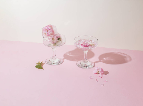 Zwei Gläser mit Eiswürfeln und rosa Blütenblättern auf rosa Hintergrund. Sonniges Tageskonzept - Foto, Bild