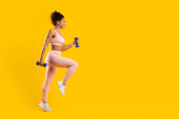 Zdeterminowana afro-amerykanka zaangażowana w rygorystyczną rutynę fitness z hantlami, podkreślając siłę i ostrość, na odizolowanym żółtym tle - Zdjęcie, obraz