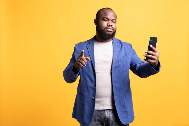 Ερεθισμένος Αφροαμερικάνος που τσακωνόταν με φίλους κατά τη διάρκεια τηλεδιάσκεψης χρησιμοποιώντας κινητό τηλέφωνο, απομονωμένος στο στούντιο. Εξοργισμένο άτομο που τσακώνεται με τους φίλους του κατά τη διάρκεια βίντεο στο διαδίκτυο - Φωτογραφία, εικόνα