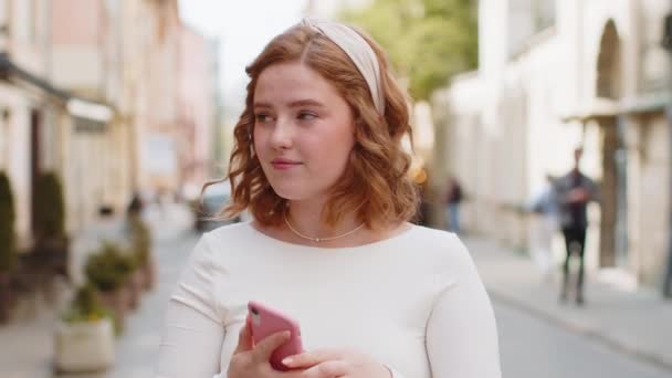 Usmívající se šťastná zrzka mladá žena pomocí smartphone psaní SMS e-mailových zpráv procházení internetové aplikace, dokončovací práce, při pohledu na kameru venku. Dívka procházející se po městské ulici. Životní styl města. - Záběry, video