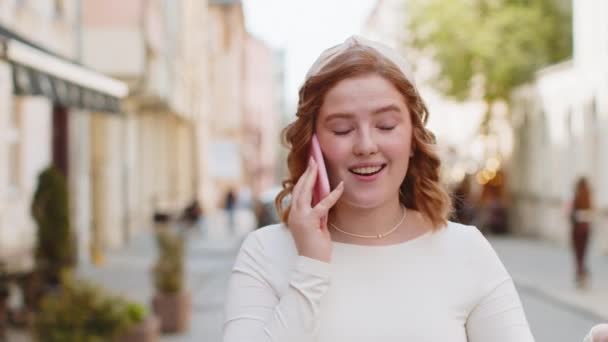 Phone call, good news, gossip. Happy surprised redhead young woman in pleasant conversation on smartphone, enjoying talking with friend outdoors. Girl walking in urban city street. Town lifestyles - Footage, Video
