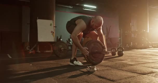 Vue latérale en temps réel d'un mâle adulte chauve fort debout dans des baskets sur le sol ajoutant une plaque de poids et un serrage avec des vis dans la salle de gym à la lumière du jour - Séquence, vidéo