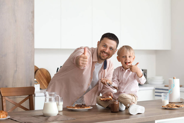 Mały chłopiec z ojcem jedzący czekoladowe tosty, pijący mleko i pokazujący gest kciuka w kuchni - Zdjęcie, obraz
