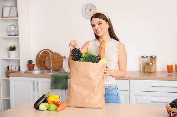 Mutfaktaki masada elinde bir torba taze sebze ve meyve olan genç bir kadın. - Fotoğraf, Görsel