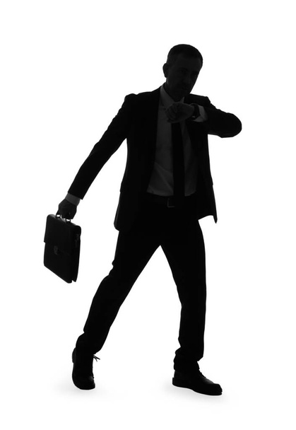 Силуэт зрелого бизнесмена с портфелем, смотрящего на наручные часы на белом фоне - Фото, изображение