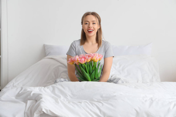 Yatak odasında bir buket lale çiçeğiyle rahat bir yatakta oturan mutlu genç kadın. - Fotoğraf, Görsel