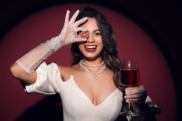Beautiful young African-American woman with glass of wine and poker chips on dark red background - Photo, Image