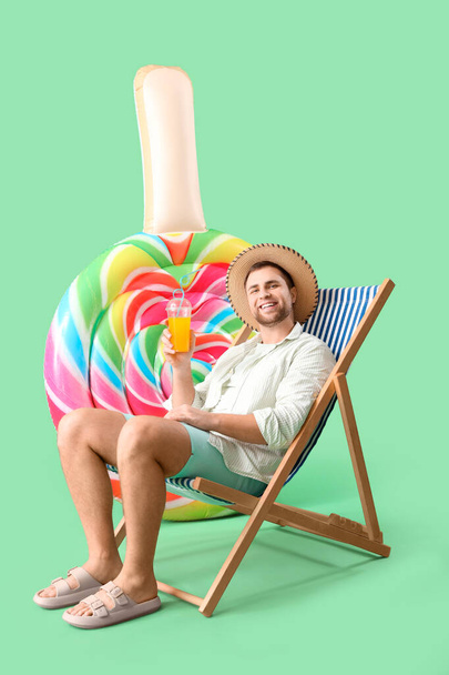 Young man with inflatable mattress in shape of candy sitting on deckchair against green background - Photo, Image