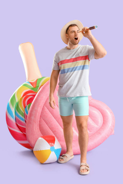 Retrato de joven sorprendido con colchones inflables mirando a través de spyglass en fondo lila - Foto, Imagen