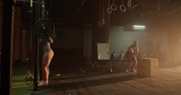 Real time side view of strong male jumping on to block from ground and later exercise with kettlebell during training with female athlete lifting heavy barbell and switching to burpee - Záběry, video