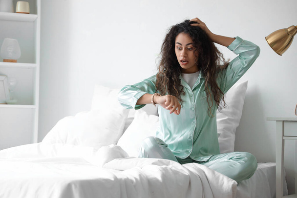 Overslept young African-American woman looking at wristwatch in bedroom - Foto, Imagen