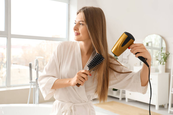 Όμορφη νεαρή γυναίκα στεγνώνει τα μαλλιά της με στρογγυλό πινέλο στο μπάνιο - Φωτογραφία, εικόνα