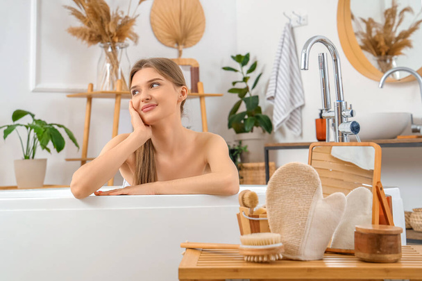 Молода жінка у ванні з масажною рукавичкою та приладдям для ванни на столі вдома - Фото, зображення